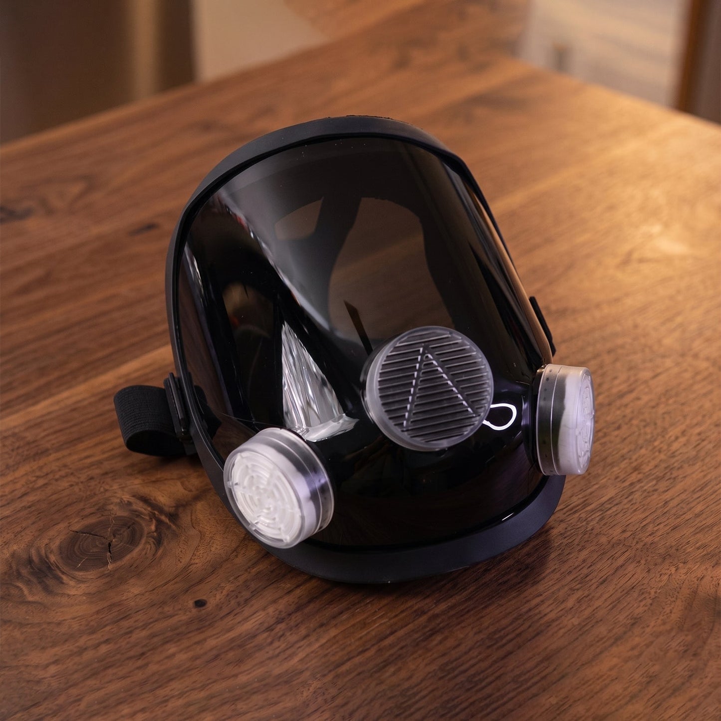 Black full face respirator mask with clear visor and two white filters resting on a wooden table.