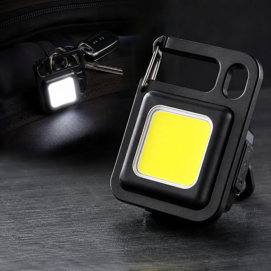 A black mini LED flashlight with a carabiner clip and a bright yellow light, next to a black bag with keys.