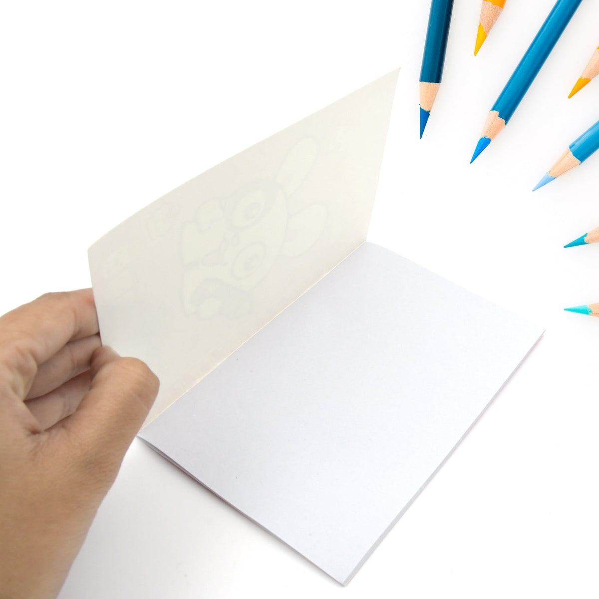 A hand holds open a white notebook with a faint bunny drawing, surrounded by blue and yellow colored pencils in a