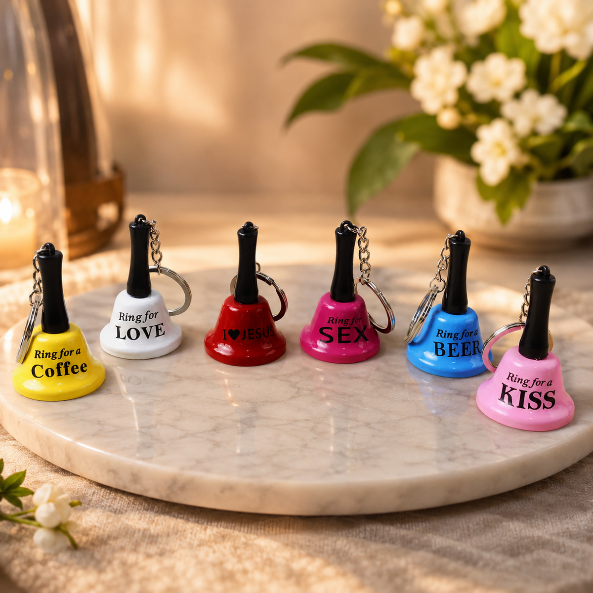 Six colorful mini hand bell keychains with various phrases on a marble surface, surrounded by soft lighting and flowers.