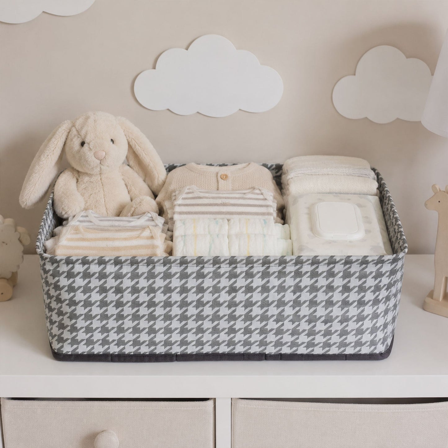 A gray and white houndstooth fabric basket holds baby clothes, diapers, wipes, and a plush bunny on a white dresser.