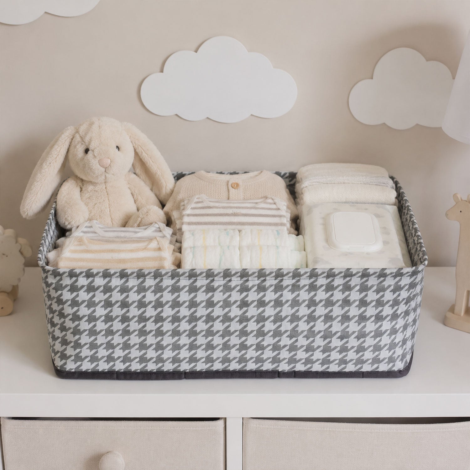 A gray and white houndstooth fabric basket holds baby clothes, diapers, wipes, and a plush bunny on a white dresser.