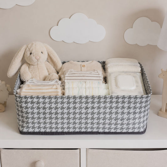 A gray and white houndstooth fabric basket holds baby clothes, diapers, wipes, and a plush bunny on a white dresser.