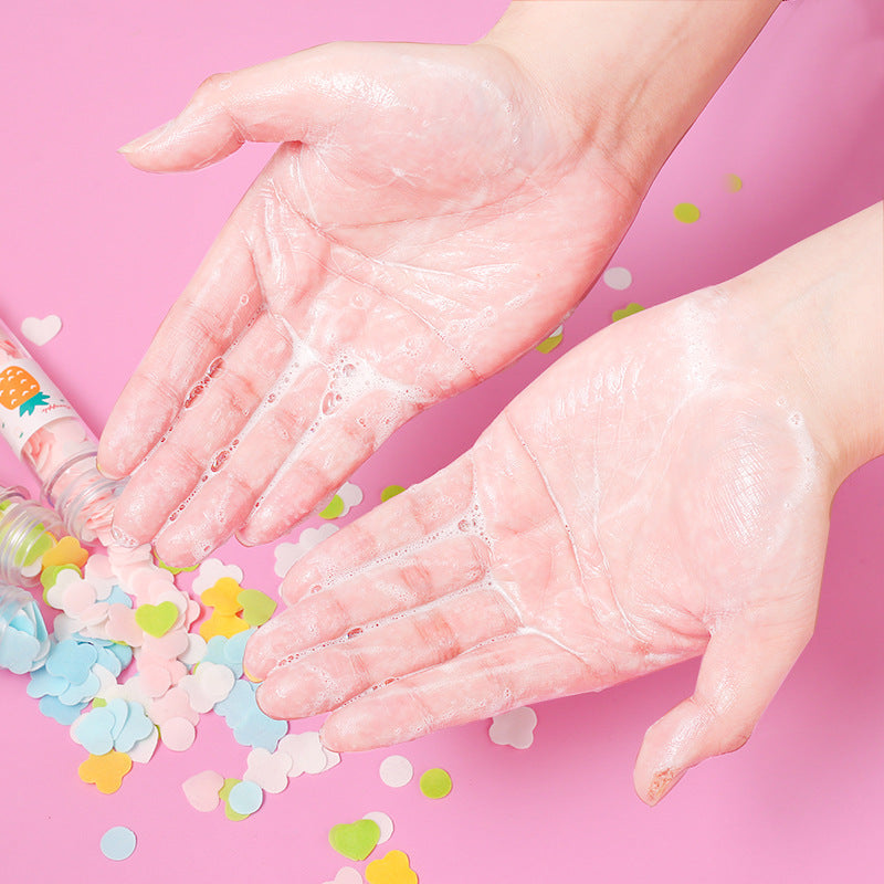 Two hands covered in white soap suds are held over a pink surface scattered with colorful flower and heart-shaped paper soap.