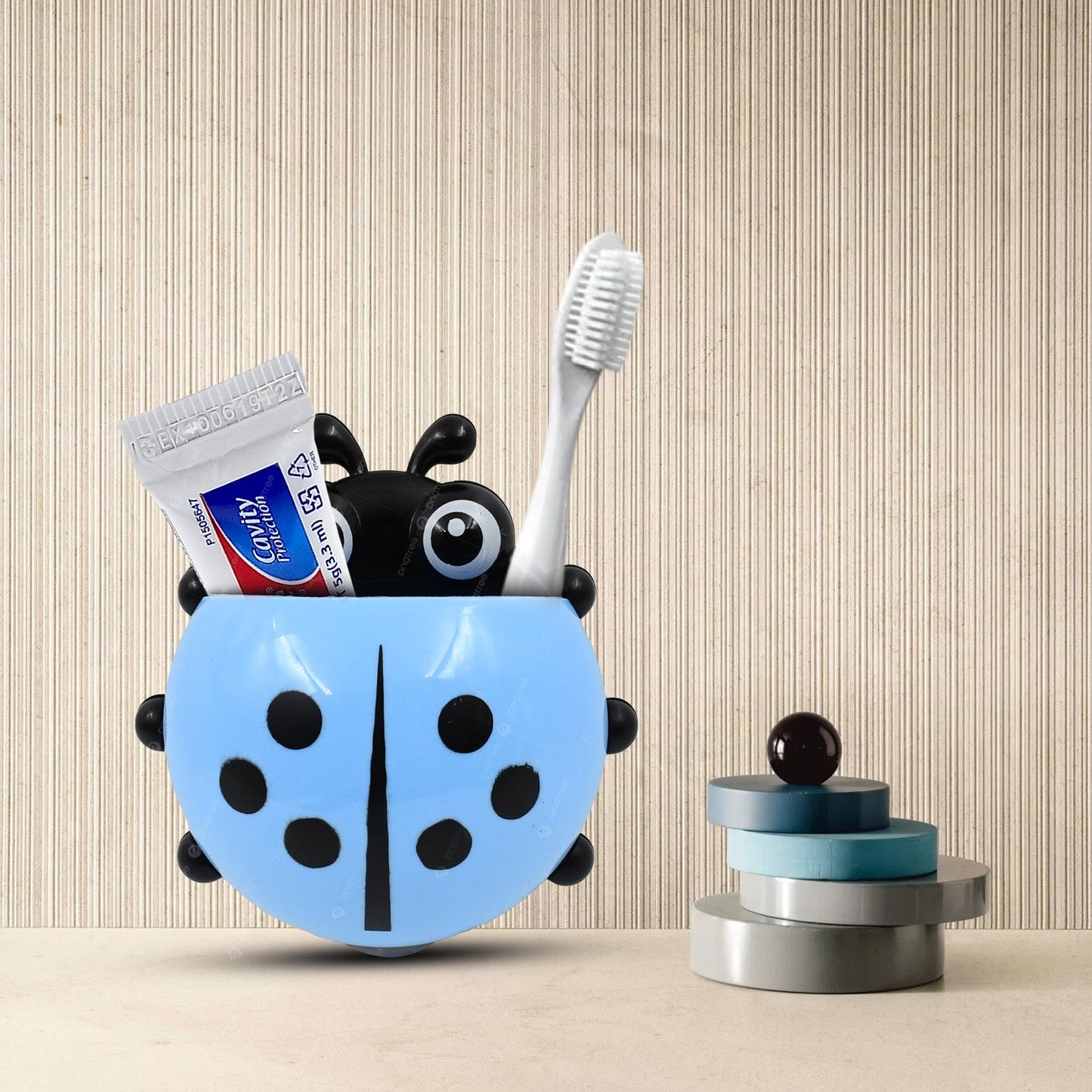 A blue ladybug holder with a toothbrush and small toothpaste tube is on a patterned wall next to decorative cylinders.
