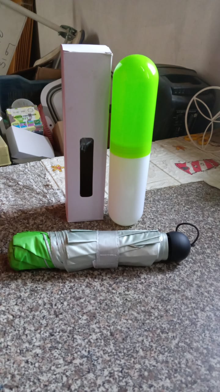 A folded green and silver umbrella, its capsule case, and a white box are on a speckled gray floor indoors.