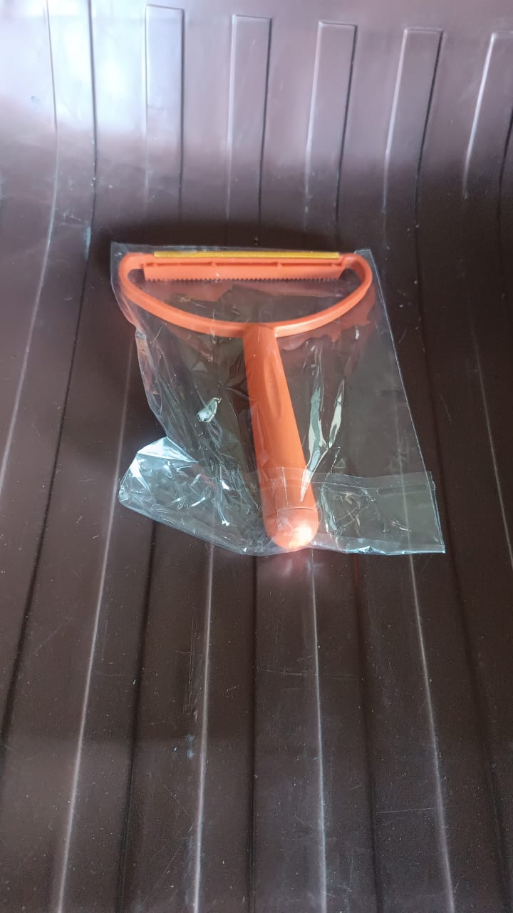 An orange lint remover with a metal shaver, sealed in clear plastic packaging, on a dark brown ribbed surface.