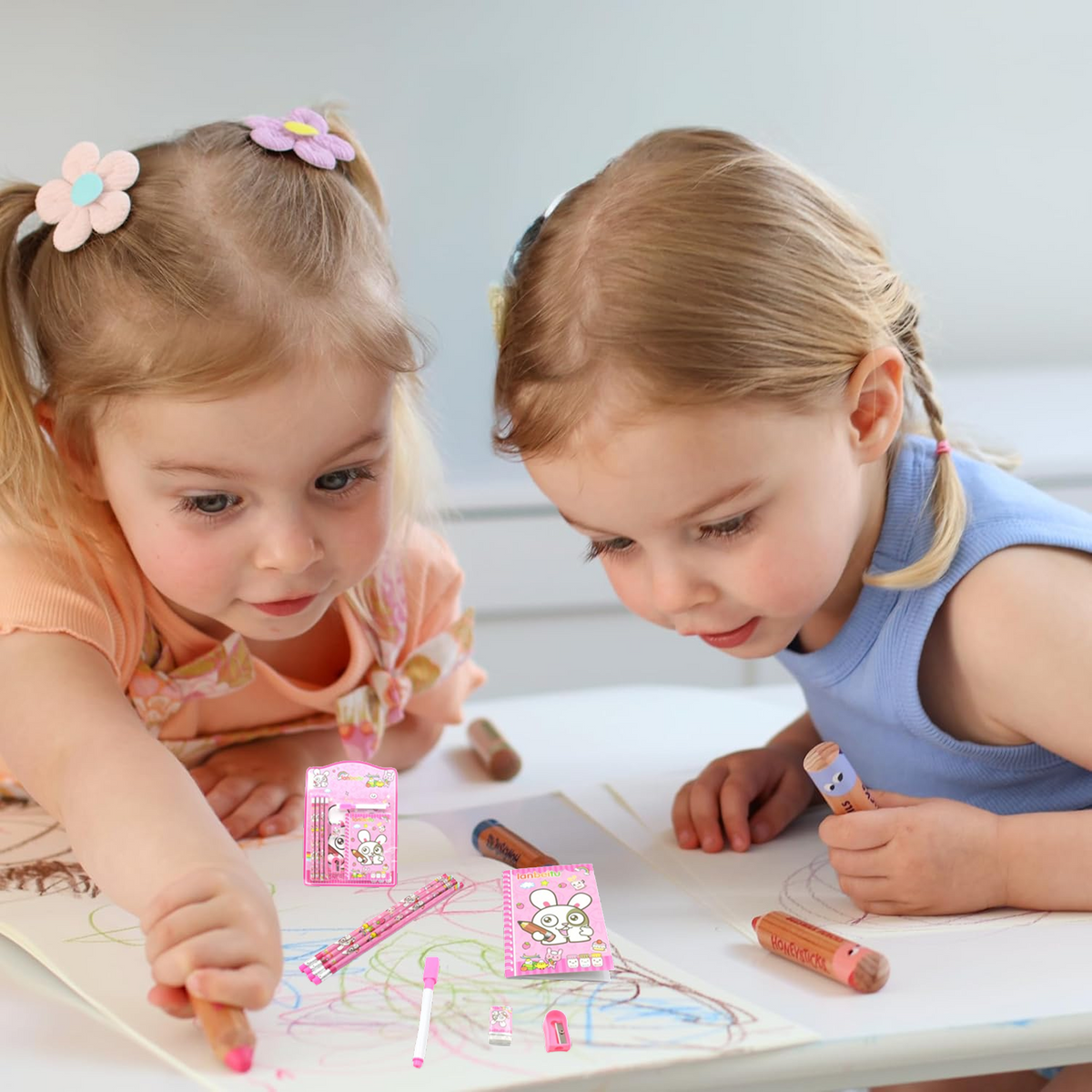 Two young girls are drawing on paper with crayons, with the pink bunny stationery set visible in the foreground.
