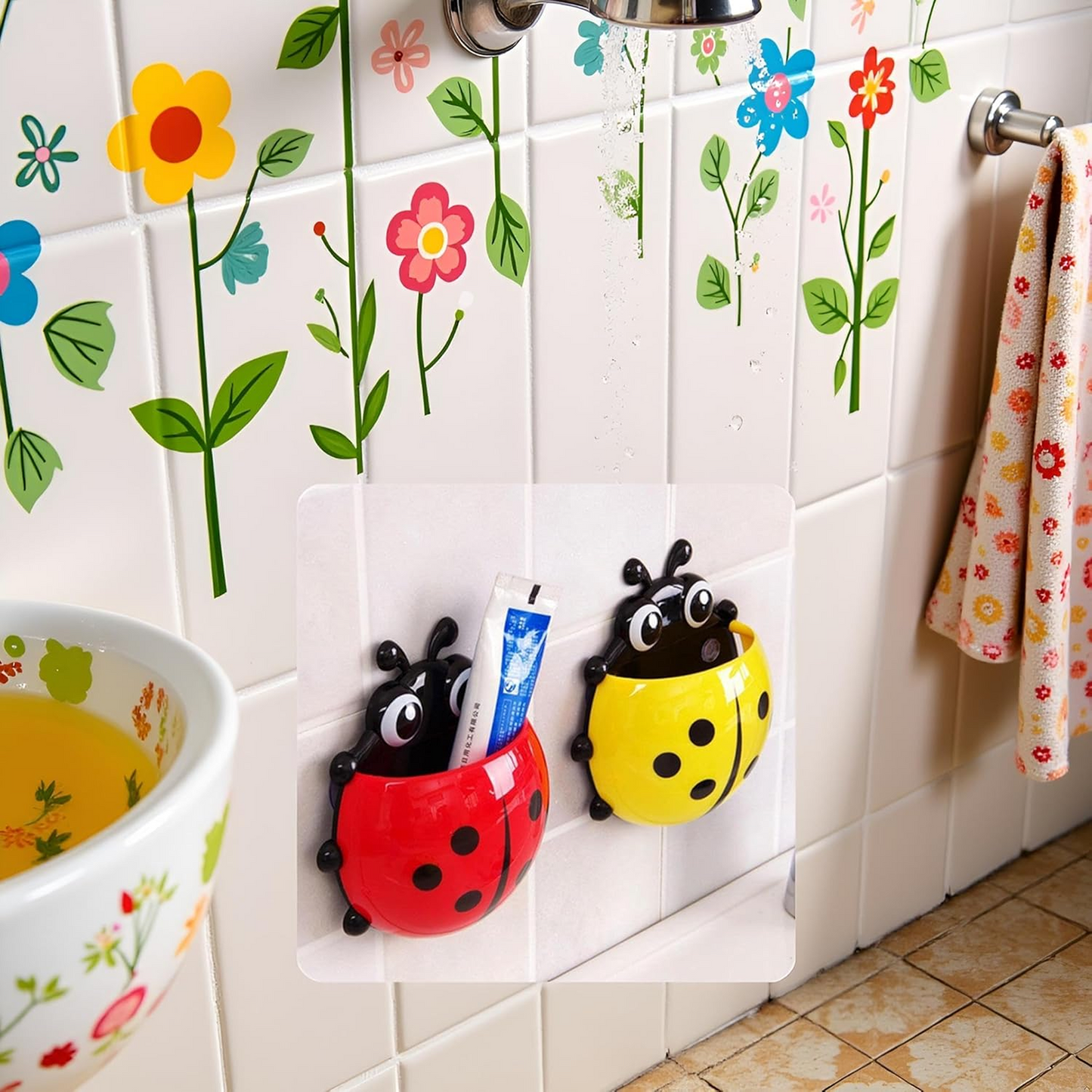 A red and a yellow ladybug holder are suctioned to a white tiled wall with floral decals in a bathroom setting.