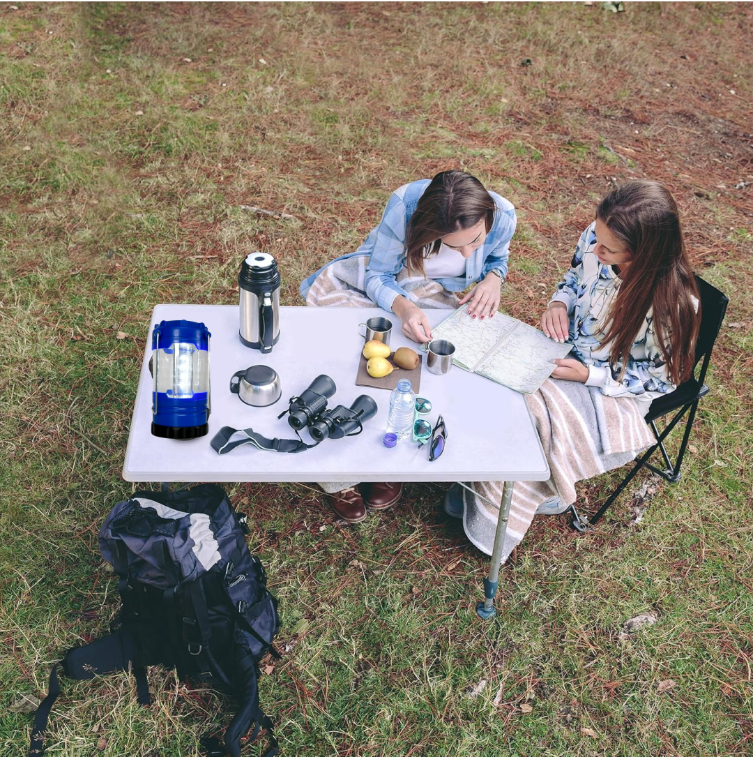 Two women sit at a white table in a grassy area, looking at a map, with a blue lantern and other camping gear.