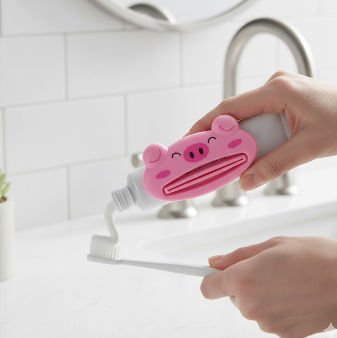 Hands using a pink pig-shaped toothpaste squeezer to dispense white toothpaste onto a white toothbrush over a bathroom sink.