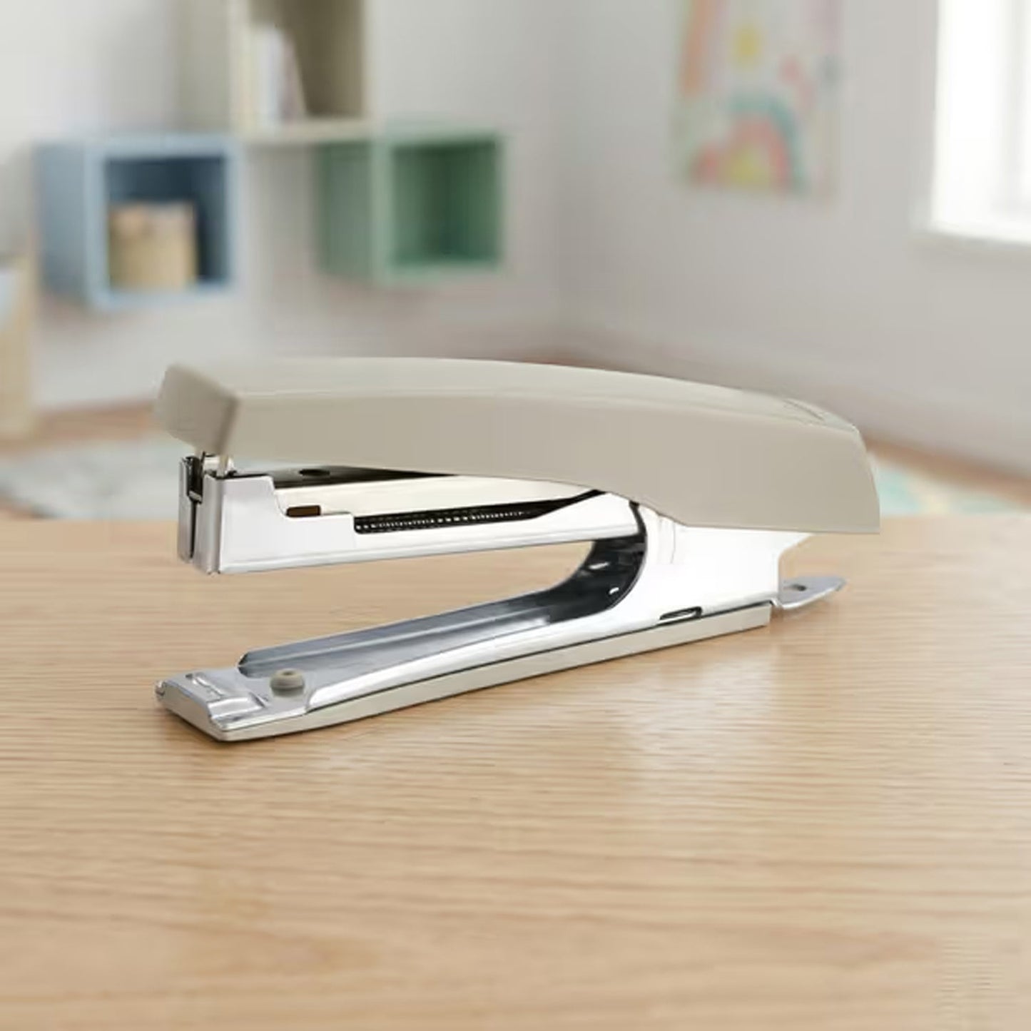 A beige and silver metal stapler sits on a light wooden desk in a room with shelves in the background.