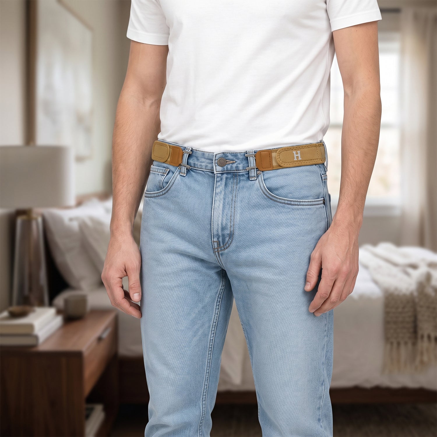 A man wearing light wash jeans and a white t-shirt, with two tan elastic belt hooks attached to his jeans, in a bedroom