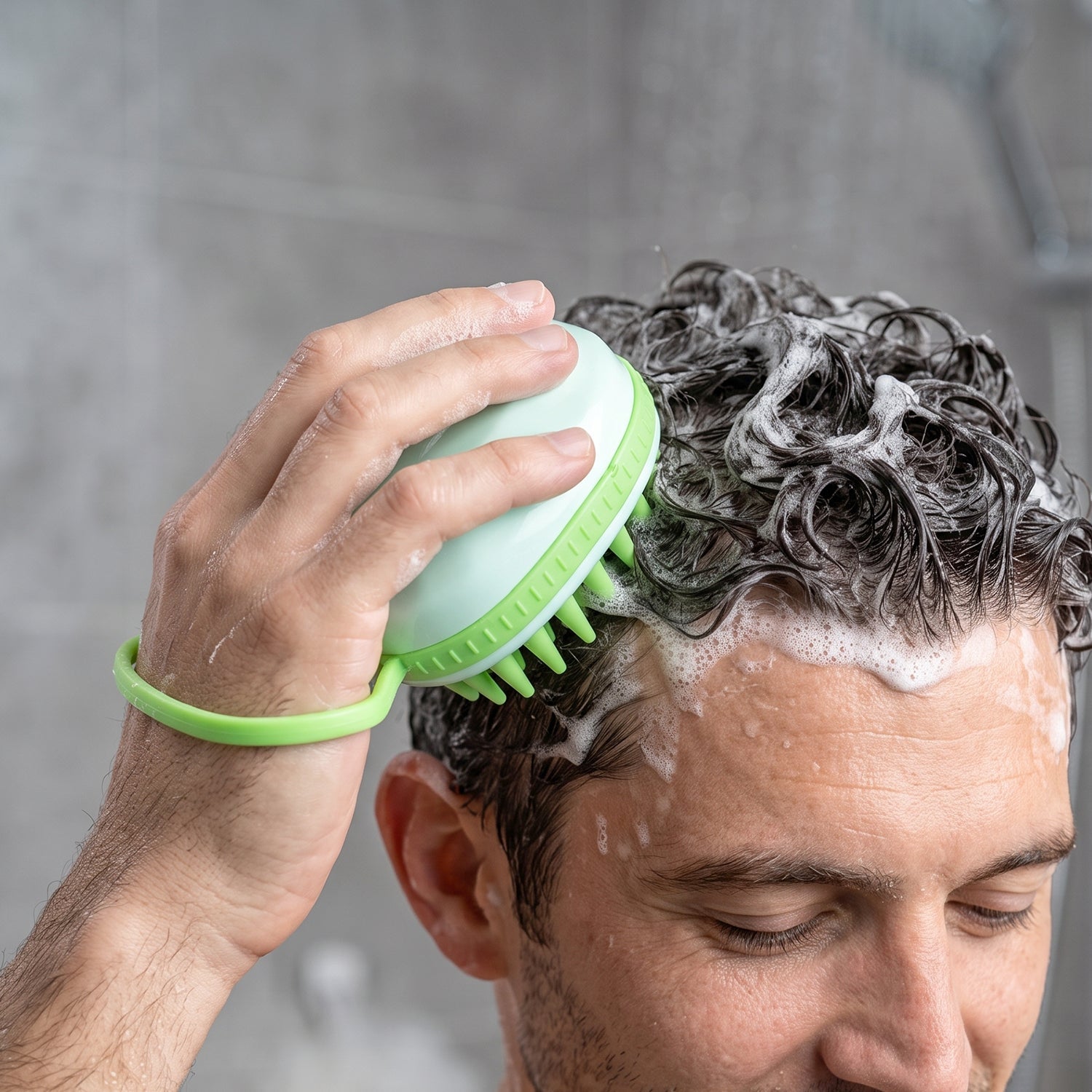 A person's hand is visible, holding a light green silicone scalp massage brush and scrubbing their dark, sudsy hair in a