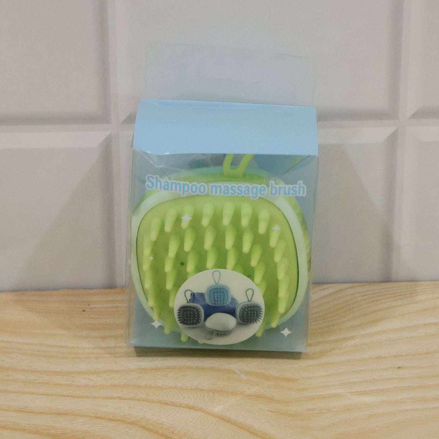 A light green silicone shampoo massage brush is packaged in a clear plastic box with a light blue top, sitting on a wooden