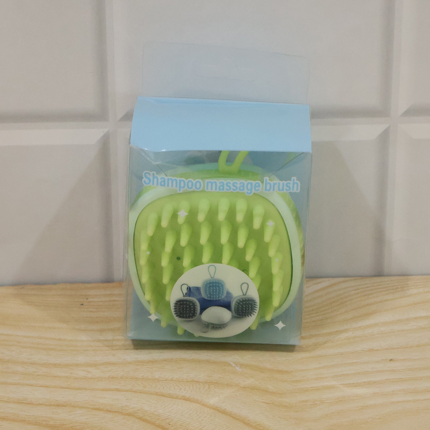 A light green silicone shampoo massage brush is packaged in a clear plastic box with a light blue top, sitting on a wooden