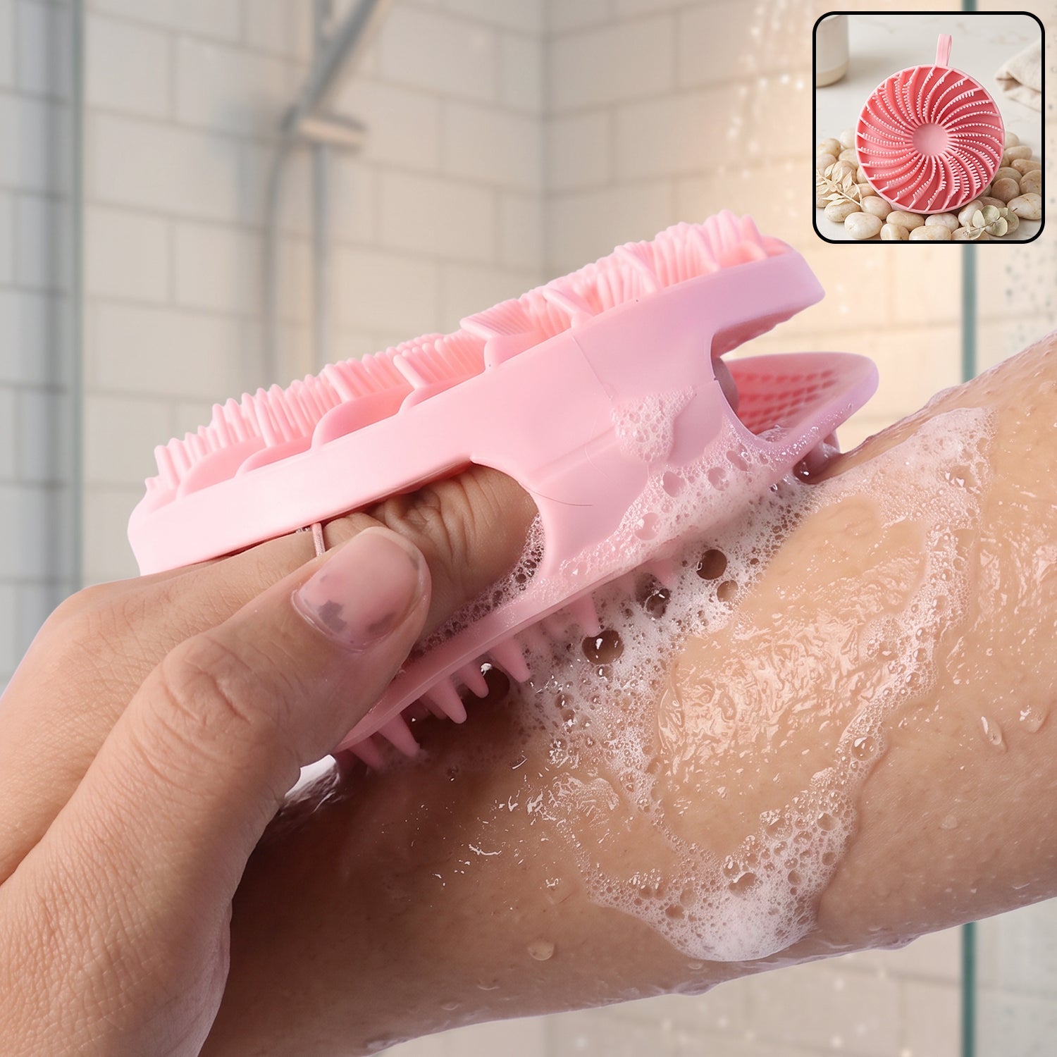 A hand holds a pink silicone body scrubber, lathering soap on an arm in a shower with a small inset image of the scrubber.
