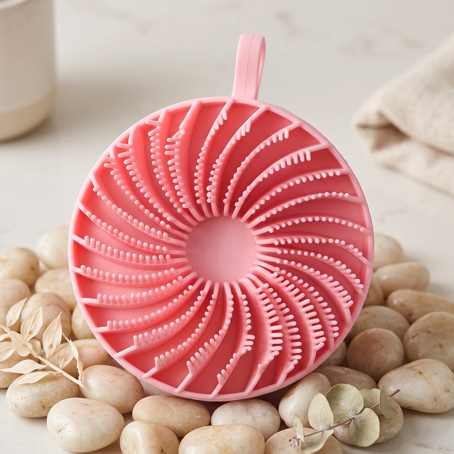 A pink silicone body scrubber with a spiral pattern of bristles rests on light-colored pebbles with a plant sprig.
