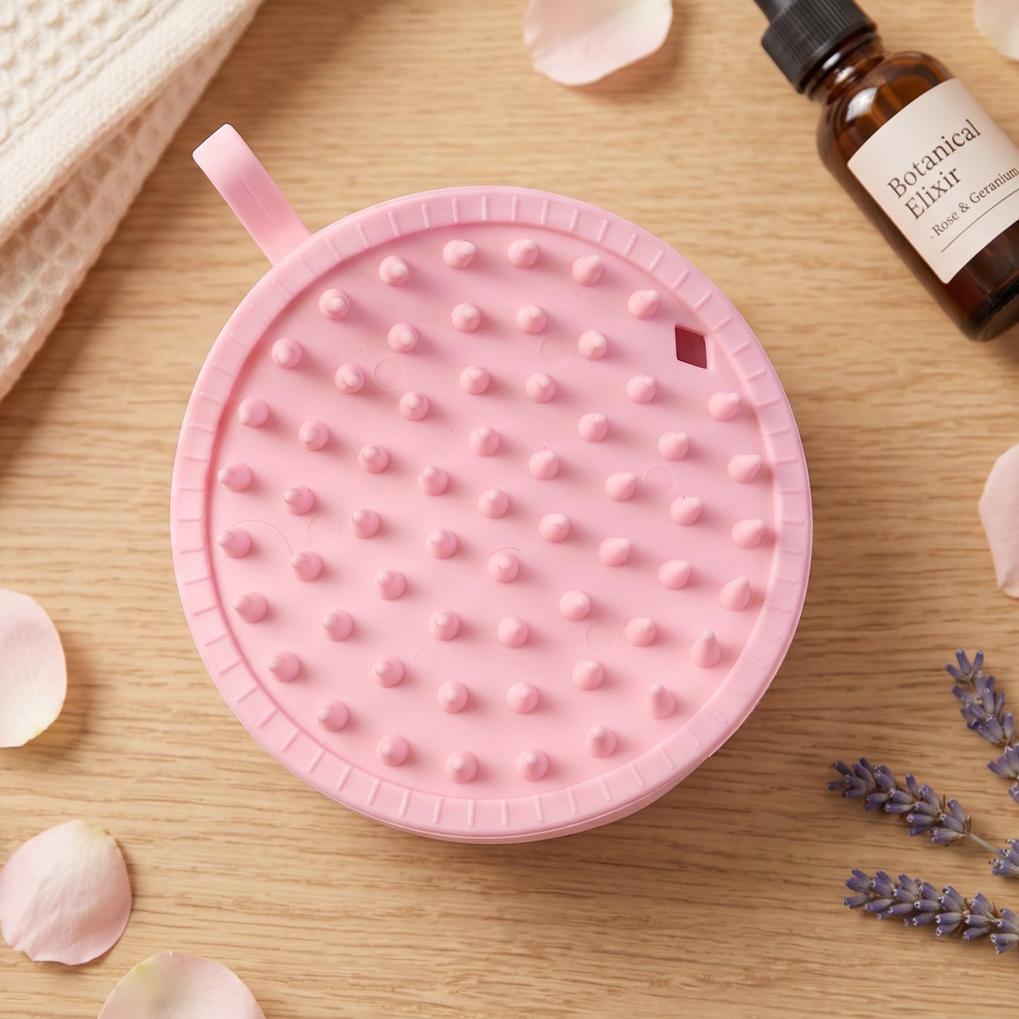 The back of a pink silicone body scrubber with round nubs is on a wooden surface with rose petals, lavender, and a bottle.