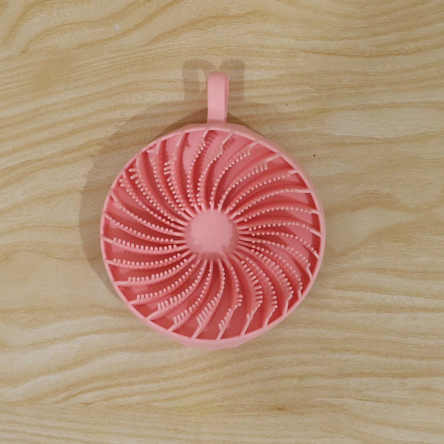 A pink silicone body scrubber with a spiral pattern of bristles and a hanging loop is centered on a light wooden surface.