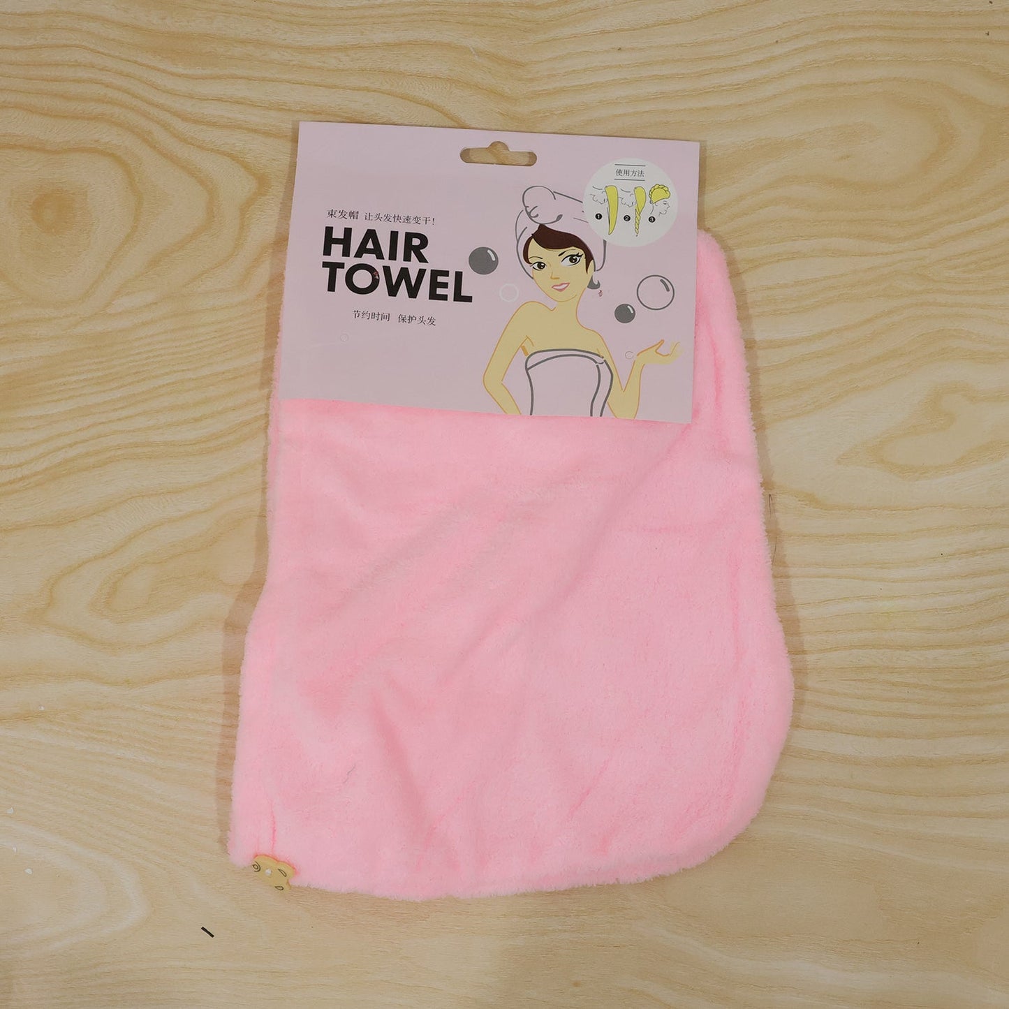 A pink microfiber hair towel is packaged with a cartoon illustration on a wooden background.