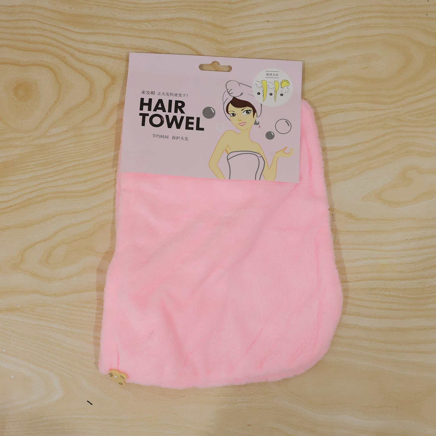 A pink microfiber hair towel is packaged with a cartoon illustration on a wooden background.