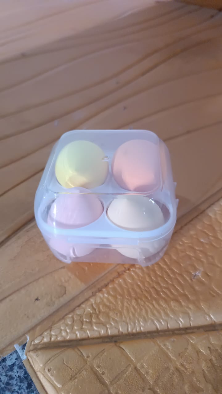 A clear plastic case containing four pastel-colored makeup sponges sits on a textured brown surface.