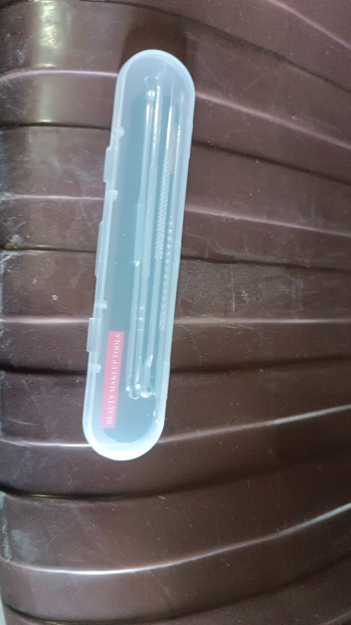 The clear plastic case containing the four stainless steel blackhead remover tools rests on a brown ribbed surface.