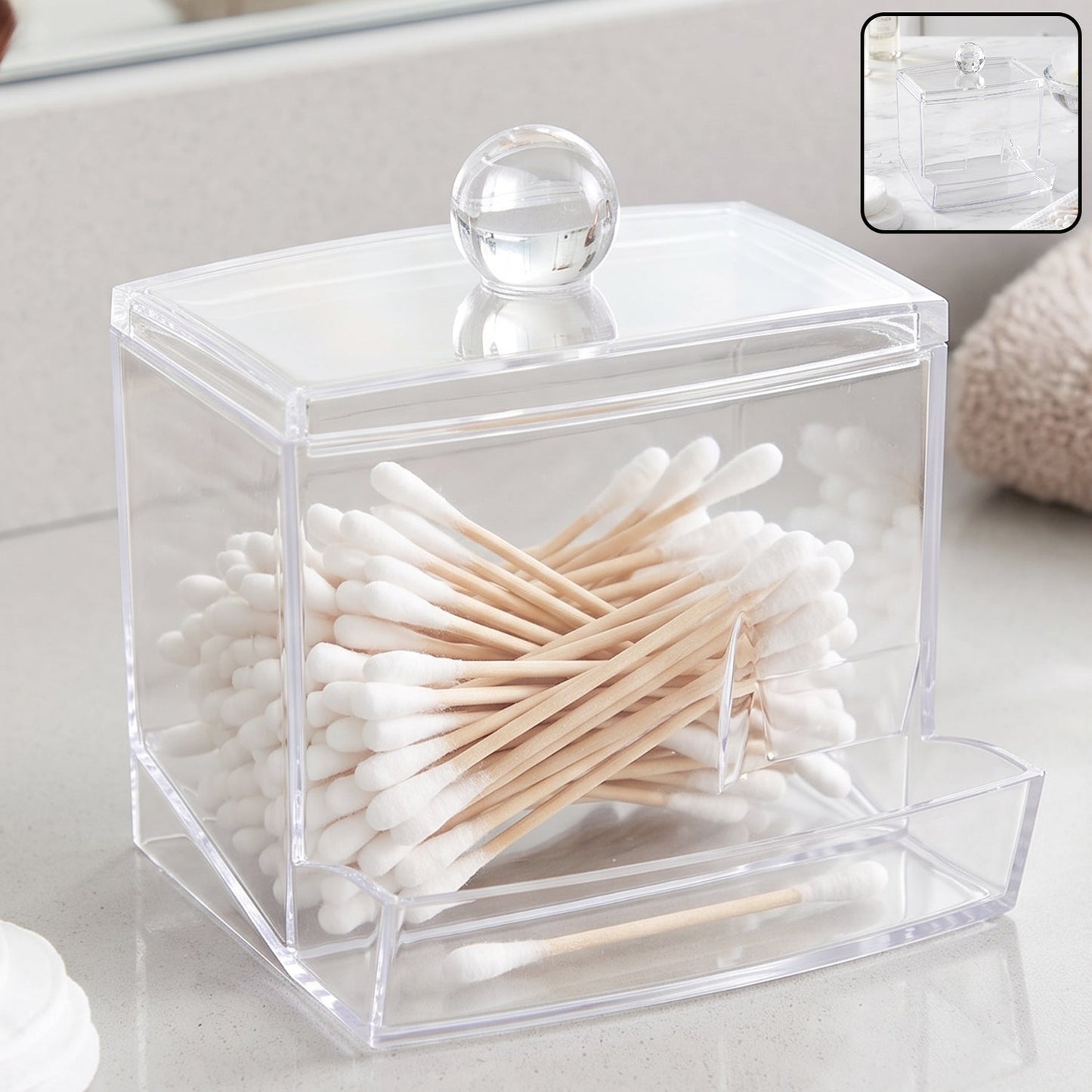 Clear acrylic cotton swab dispenser filled with cotton swabs, on a white bathroom counter.