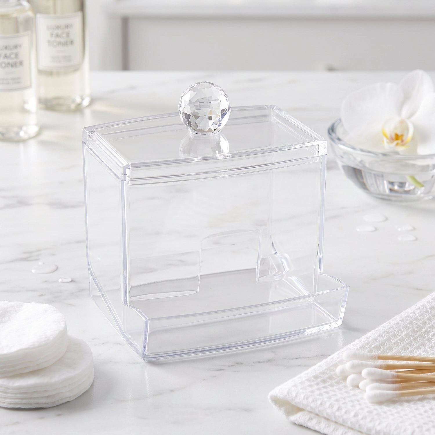 Empty clear acrylic cotton swab dispenser on a white marble counter with other toiletries.