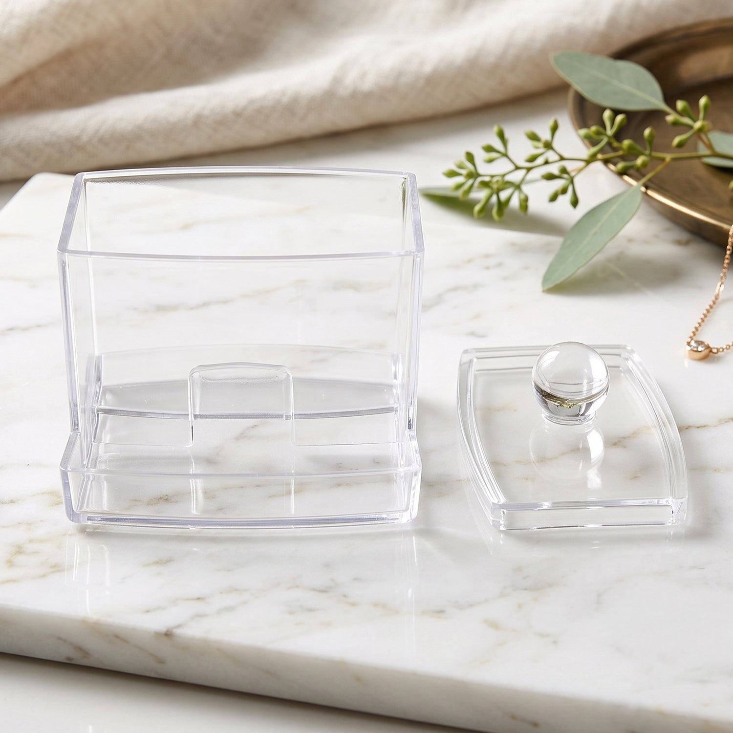 Empty clear acrylic cotton swab dispenser with its lid removed, on a white marble surface.