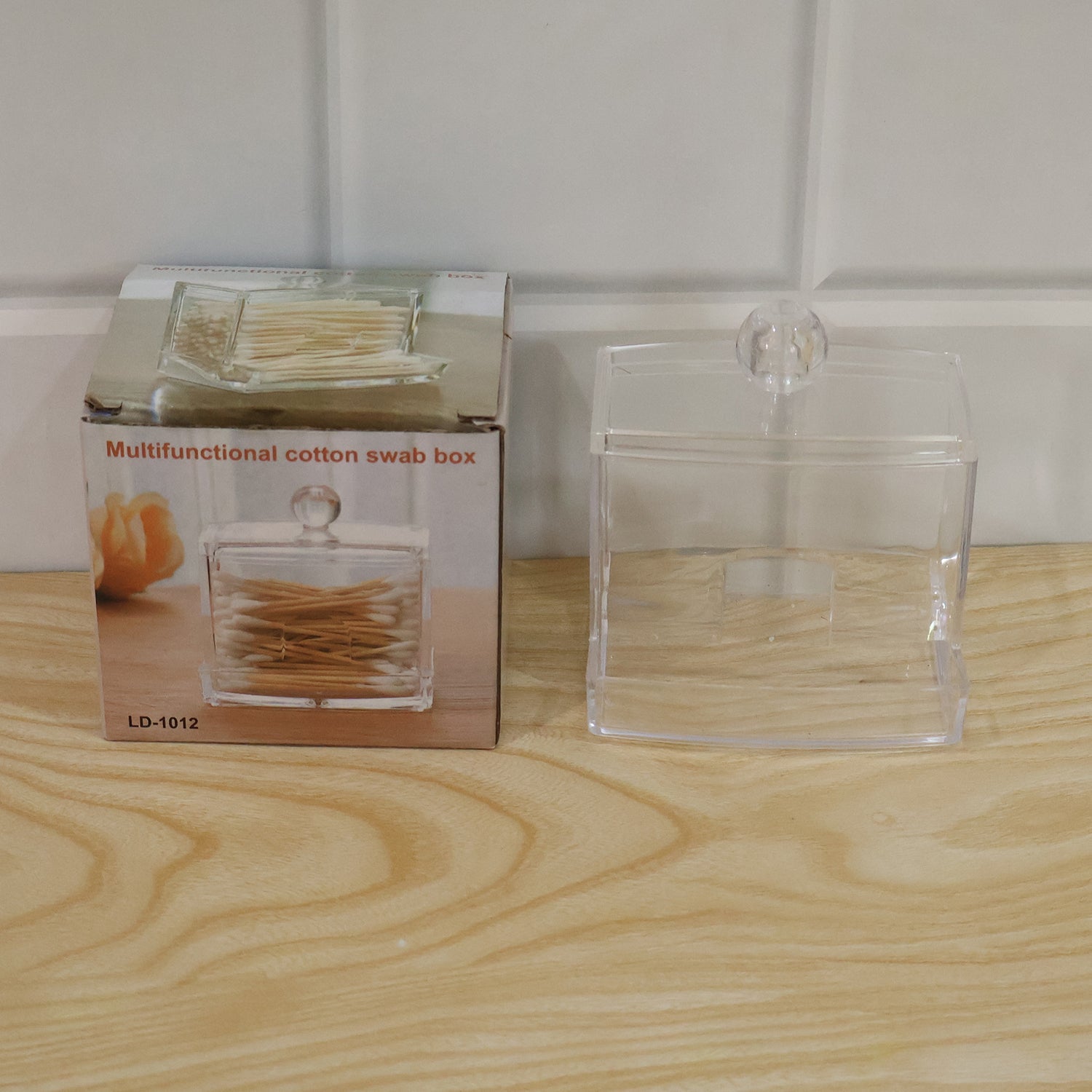 Clear acrylic cotton swab dispenser next to its product packaging box on a wooden surface.