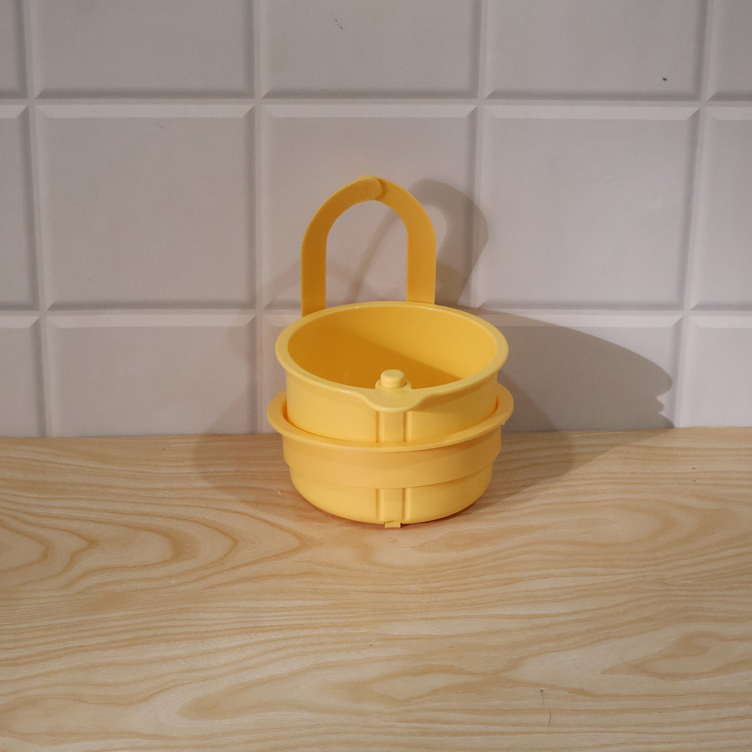 A yellow plastic hanging kitchen sink drain basket and food strainer with handle sits on a wooden counter against white