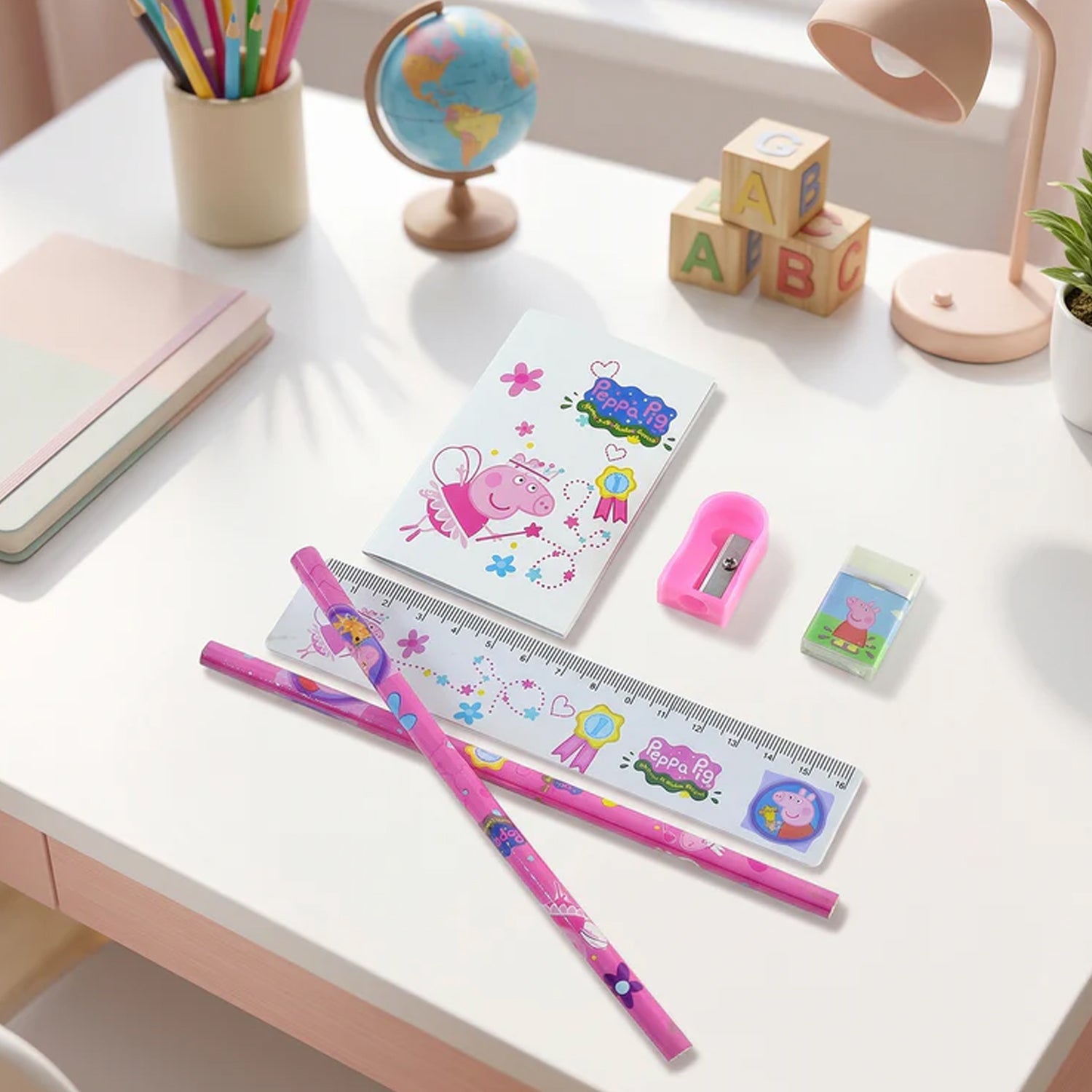 A Peppa Pig stationery set on a white desk with a globe, building blocks, and a lamp in the background.