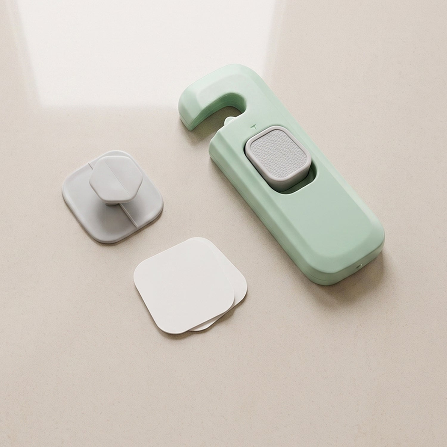 A disassembled green and grey child safety lock with two white adhesive pads on a light-colored surface.
