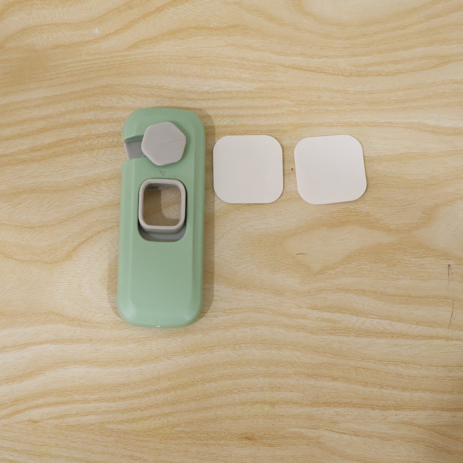 A green and grey child safety lock with two white adhesive pads on a light wooden surface.