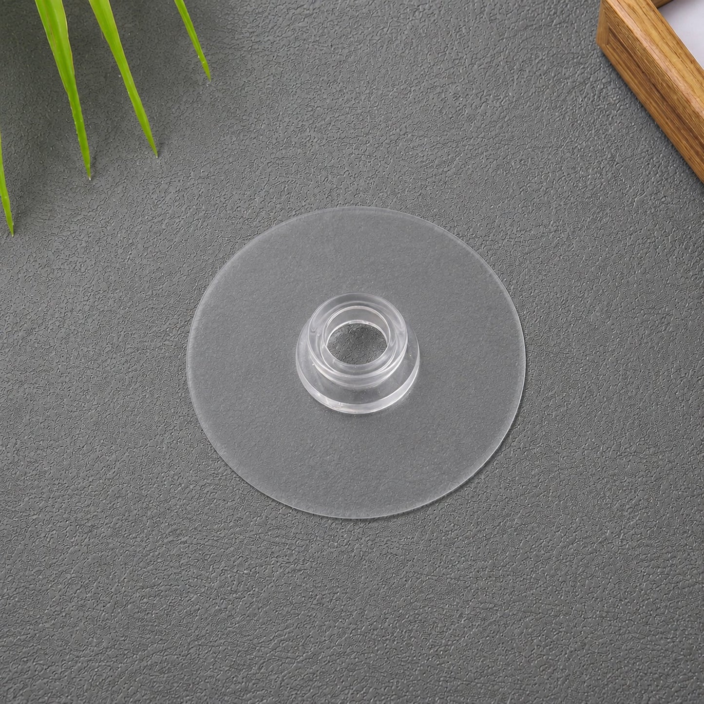 A clear plastic suction cup hook is centered on a dark grey surface, with green plant leaves and a wooden frame in the