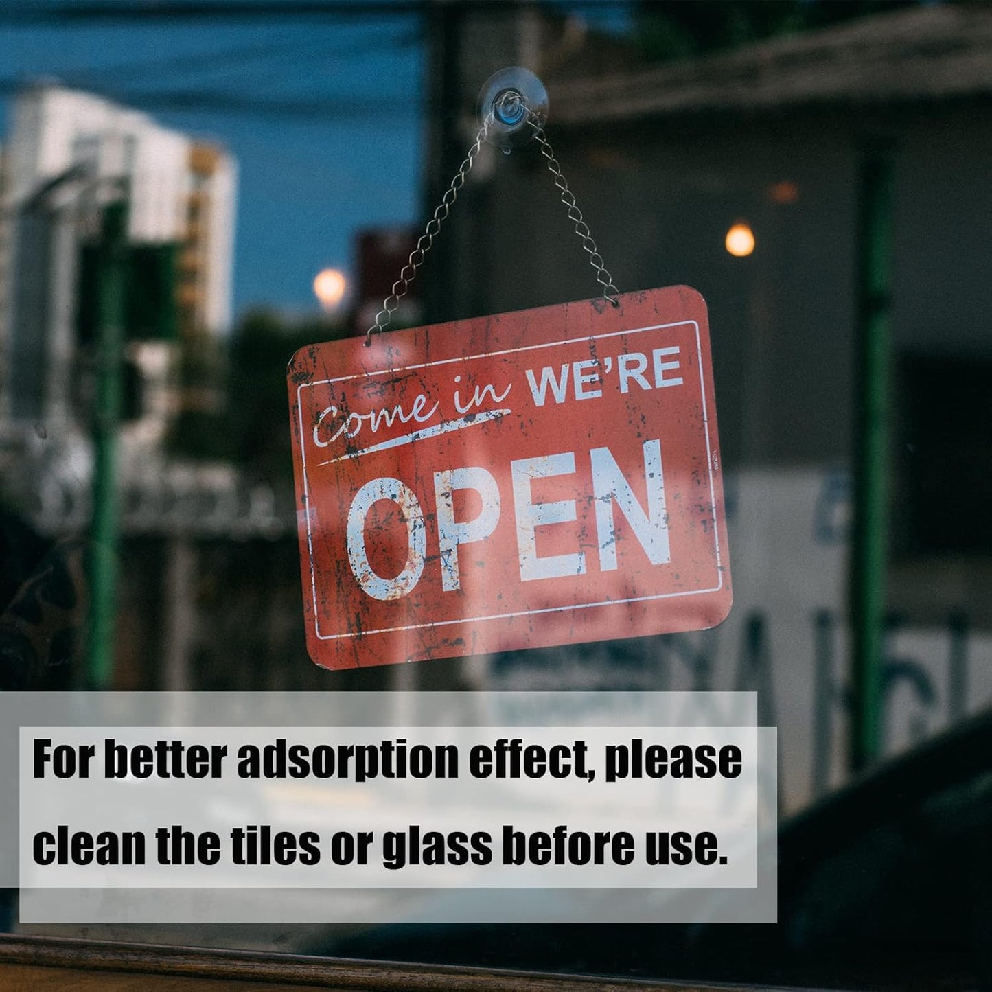 A red 'Come in We're Open' sign hangs by a chain from a suction cup hook on a glass window, with blurred buildings outside.