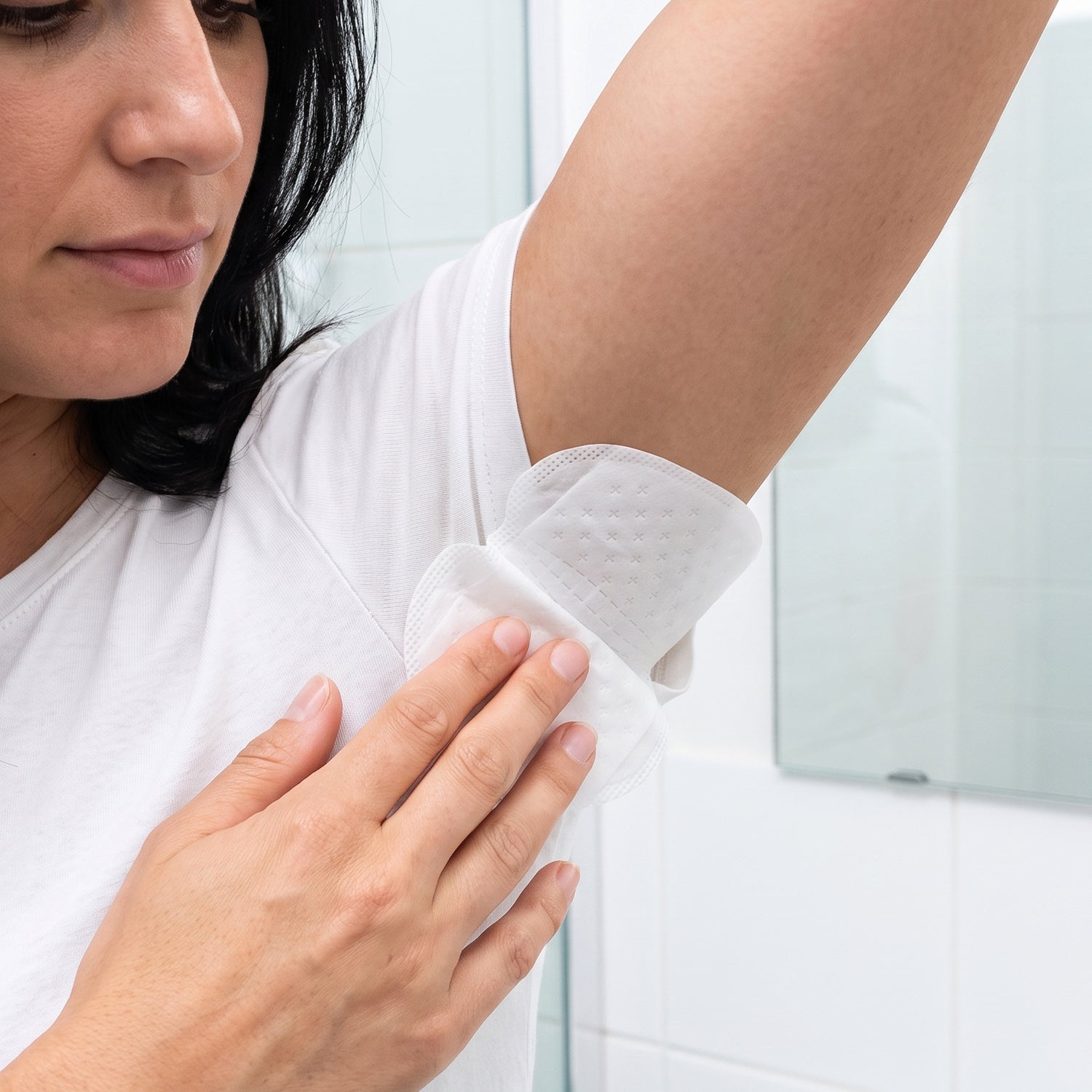 A woman in a white t-shirt applies a white, disposable underarm sweat pad to her armpit.