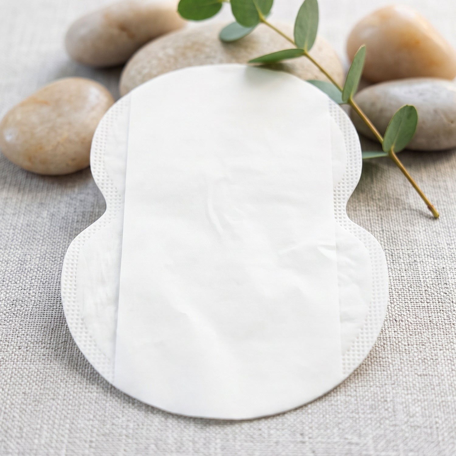 A single white, disposable underarm sweat pad with a smooth surface, surrounded by pebbles and a green sprig.