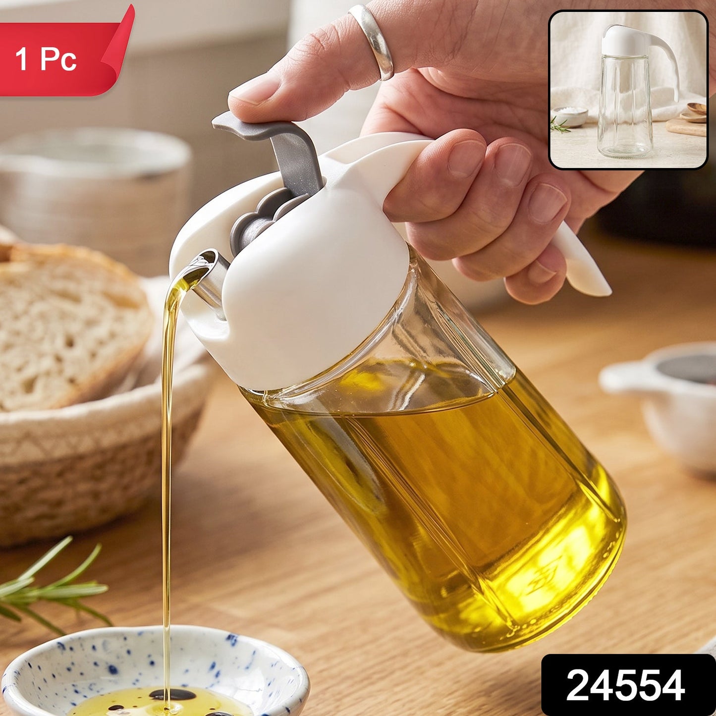 A clear glass oil dispenser with a white and gray lid is held by a hand, pouring oil into a small bowl.
