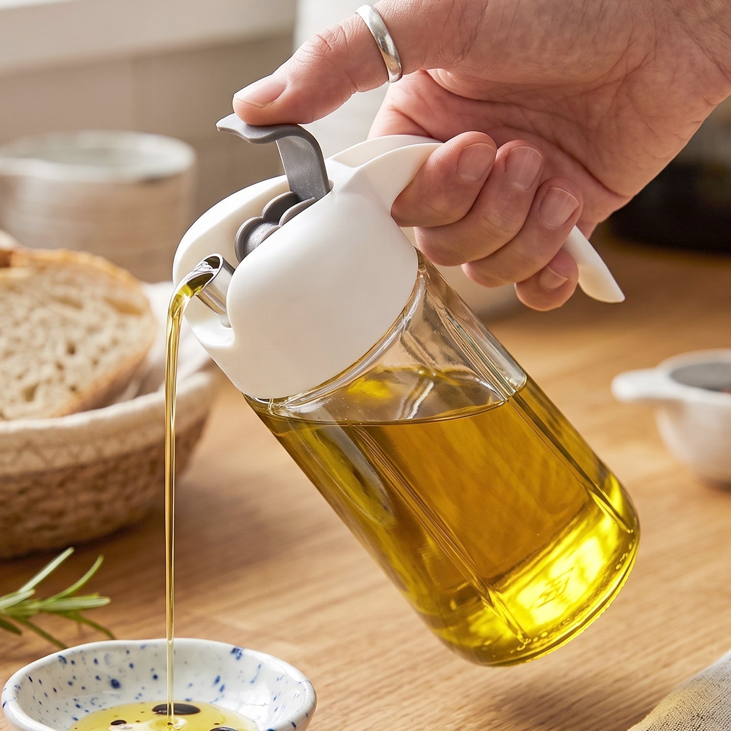 A clear glass oil dispenser with a white and gray lid is shown pouring oil into a small dish on a wooden surface.