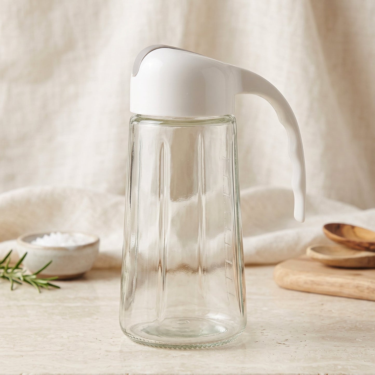 An empty clear glass oil dispenser with a white and gray lid sits on a light-colored surface with kitchen items.