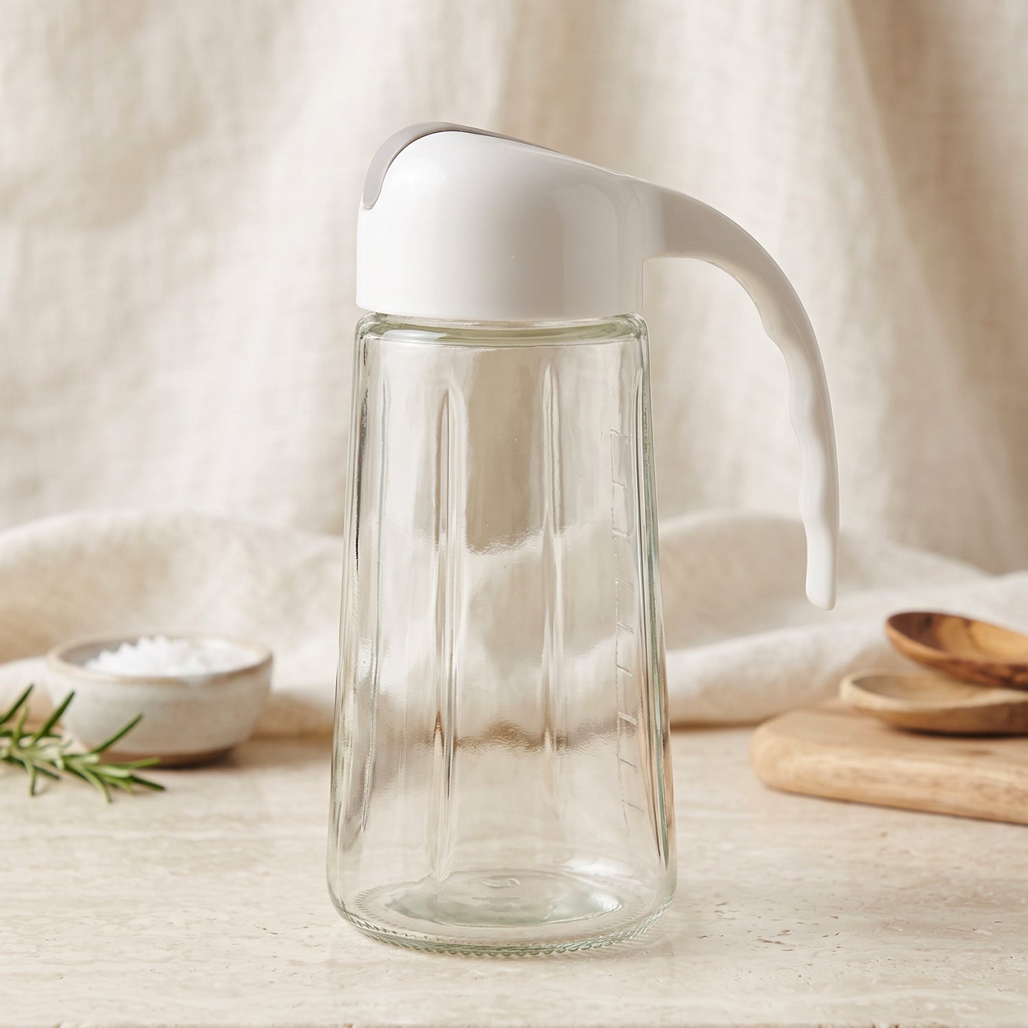 An empty clear glass oil dispenser with a white and gray lid sits on a light-colored surface with kitchen items.