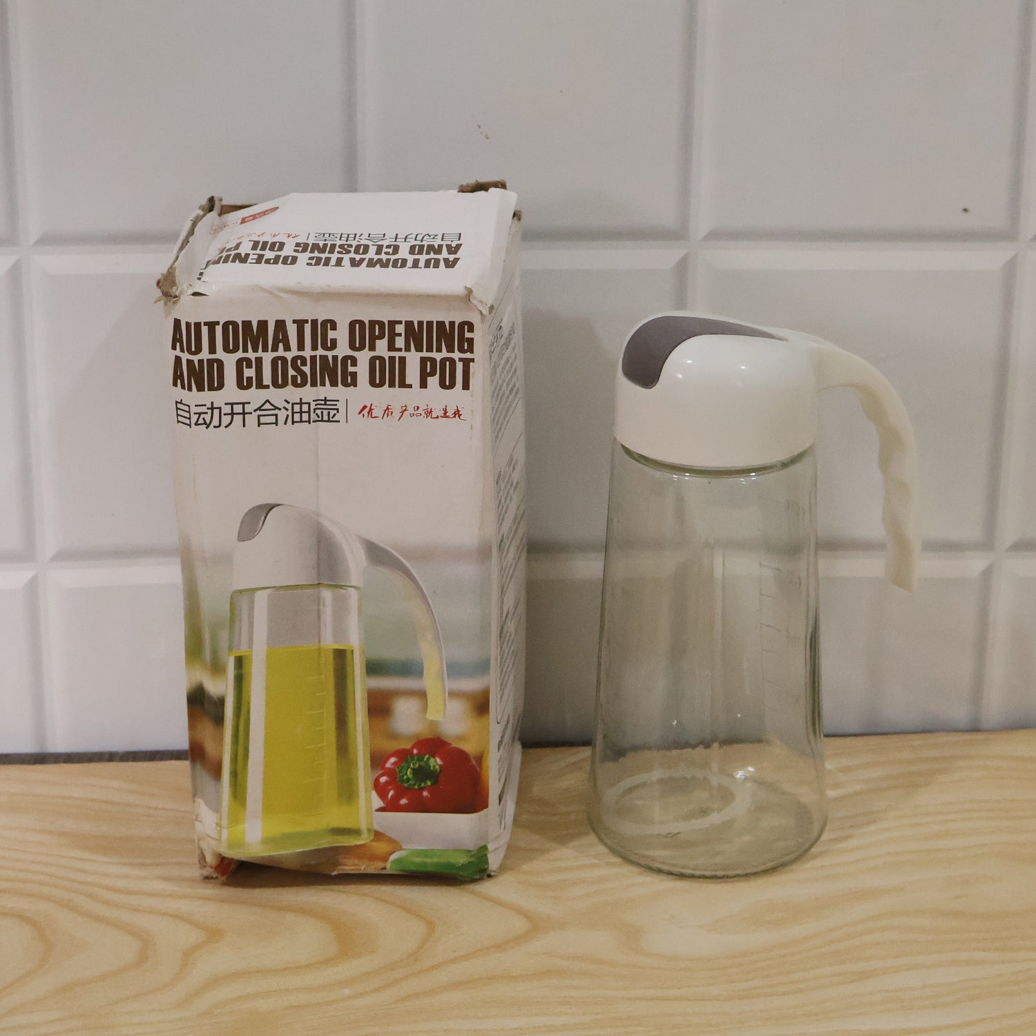 An empty clear glass oil dispenser with a white and gray lid is next to its product box on a wooden surface.
