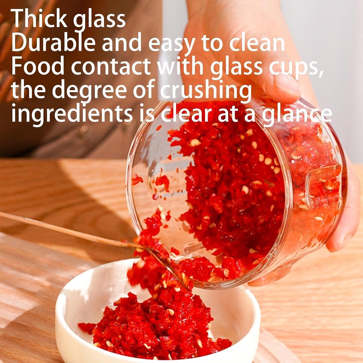 A hand pours finely chopped red chili from the clear glass bowl of the food chopper into a small white bowl.