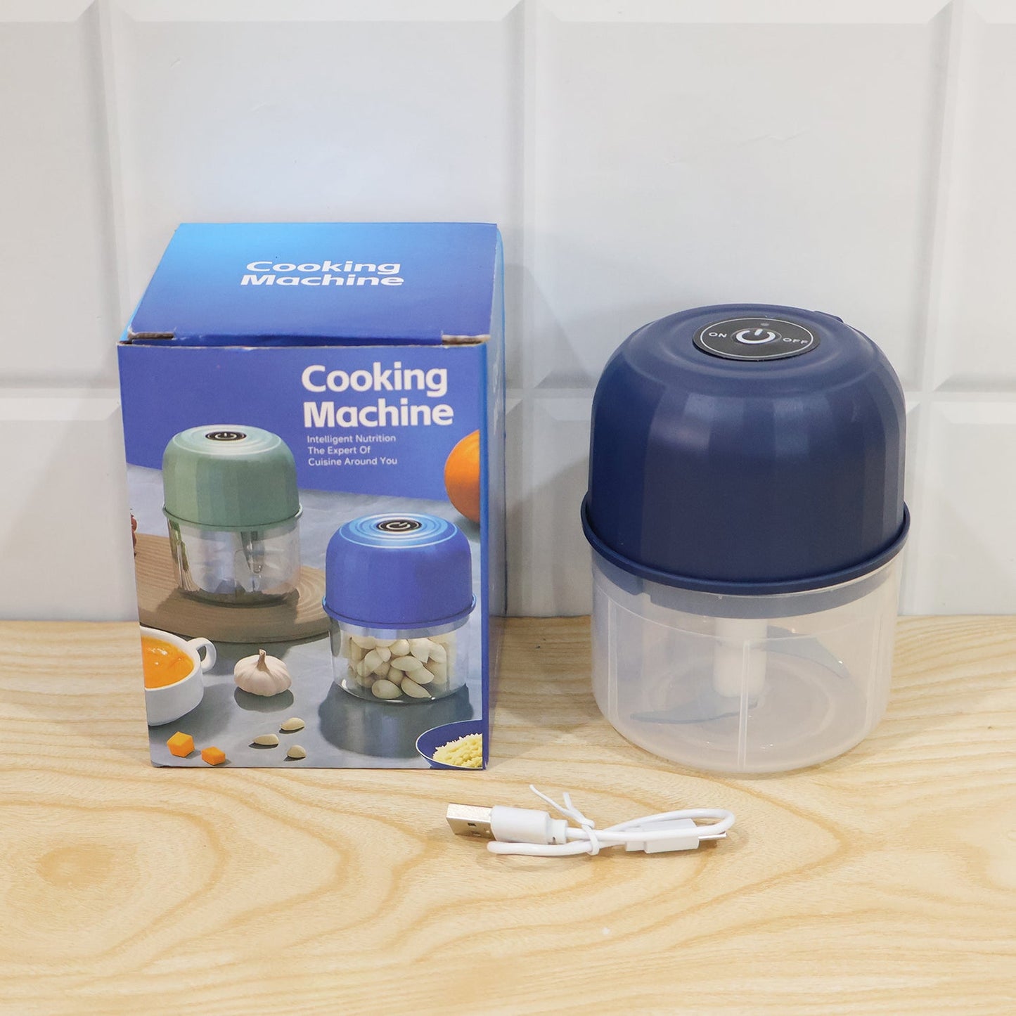 A dark blue mini food chopper, its blue box, and a white USB charging cable are arranged on a light wooden surface.