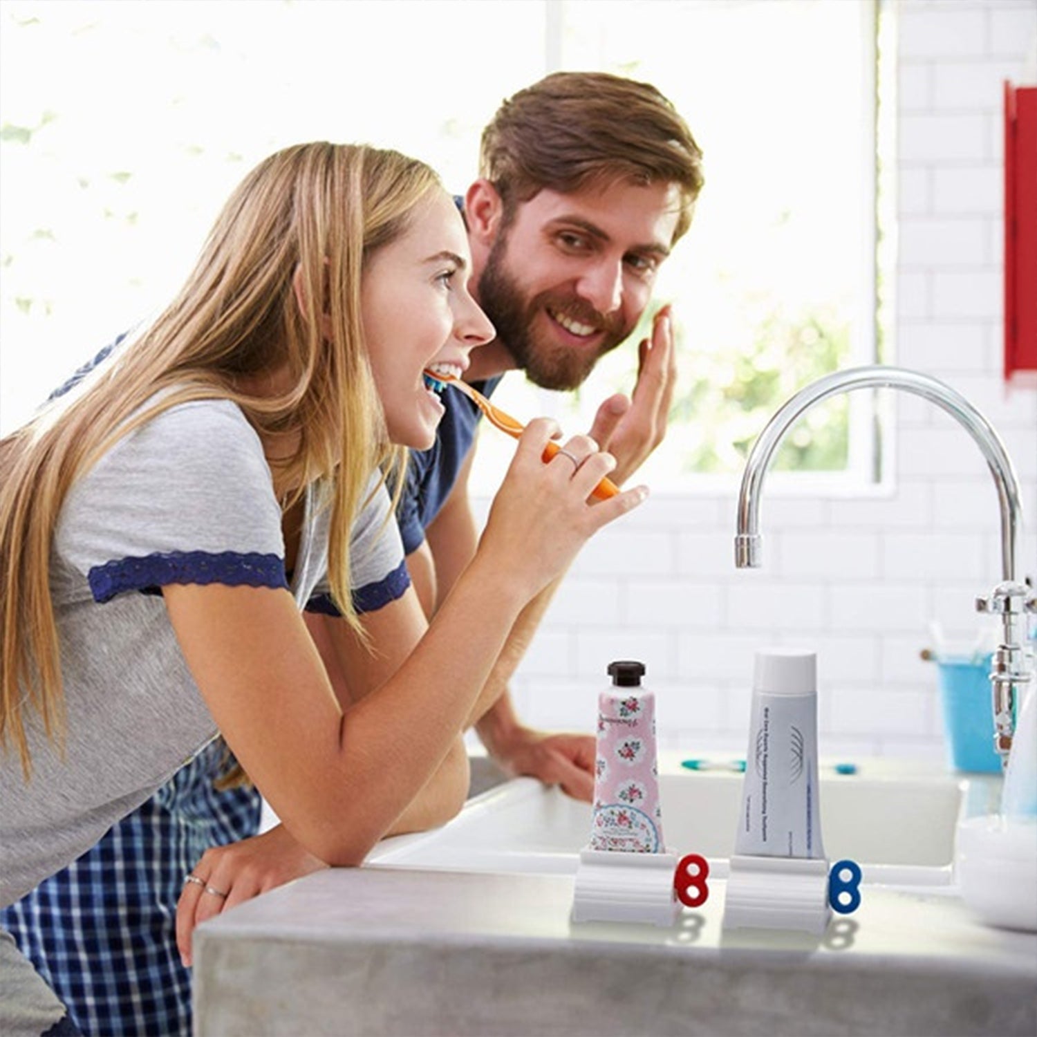 A couple brushes their teeth in a bathroom, with two tube squeezers holding toothpaste tubes on the counter.