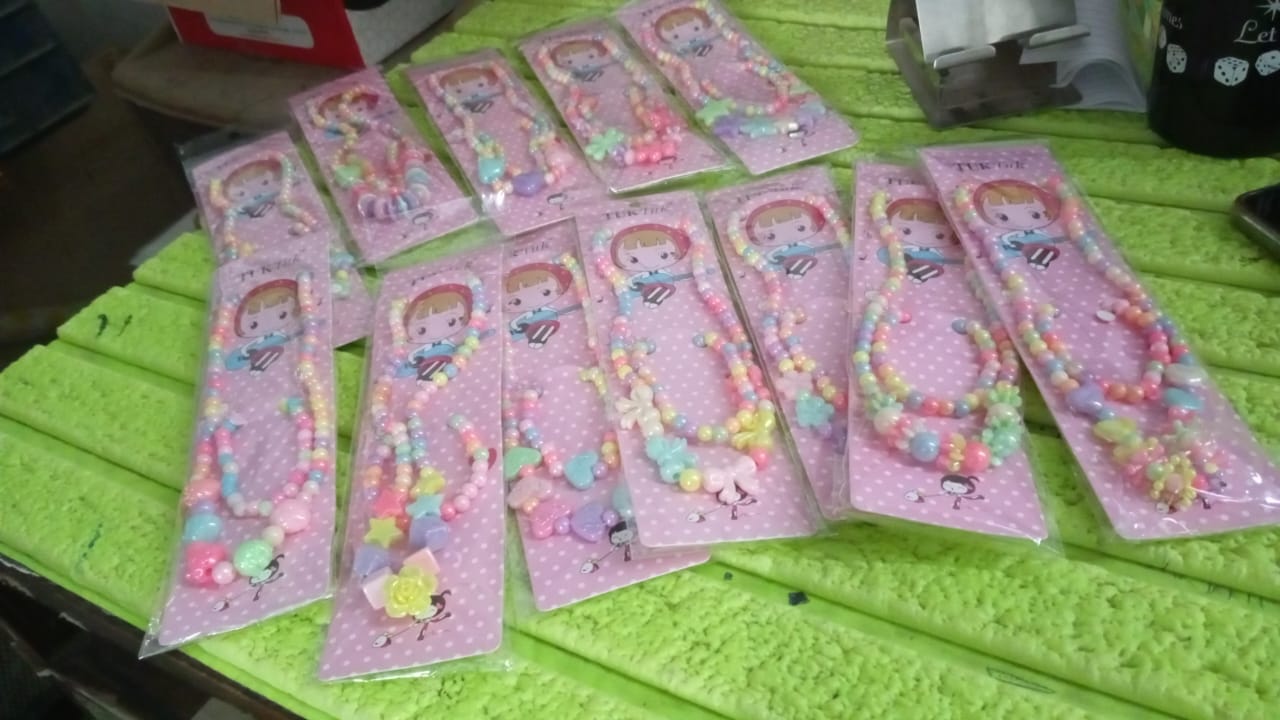 Multiple colorful beaded necklace and bracelet sets are individually packaged in pink plastic, arranged on a bright green