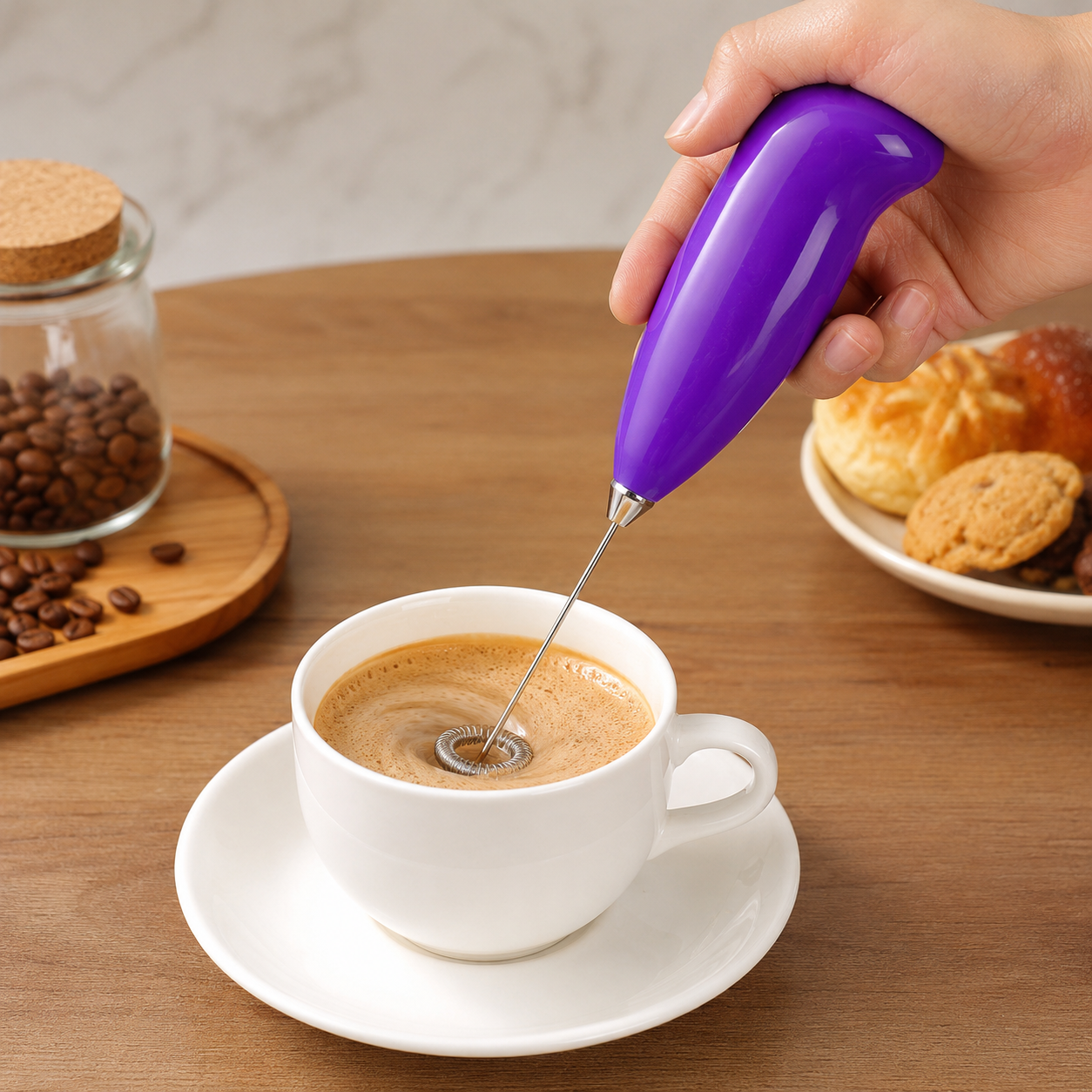 A hand holds a purple frother, whisking a cup of coffee with a frothy top, next to coffee beans and pastries.